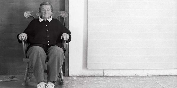 Artist Agnes Martin sitting in rocker in front of one of her paintings.