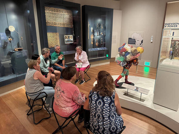 Adults seated in a museum gallery listen to a gesturing man. Nearby, a vibrant sculpture depicts a figure carrying a netted load.