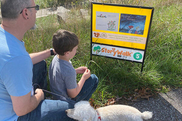 Man, boy, and white dog look at a yellow StoryWalk sign number 5 by a pond on a sunny day.