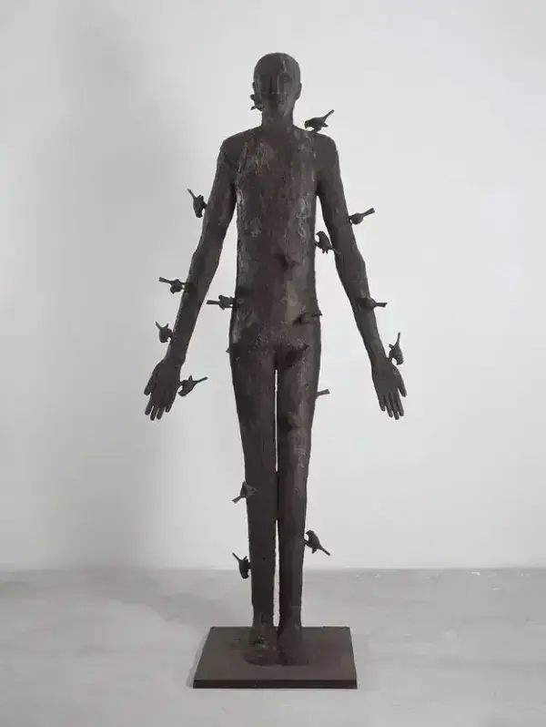Textured dark bronze sculpture of a standing human figure, with numerous small birds seemingly emerging from its body.