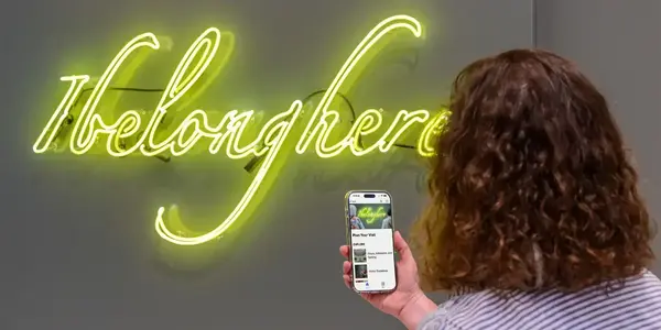 From behind, a person holds a smartphone displaying a bright yellow-green neon sign that reads 'I belong here' on a gray wall.