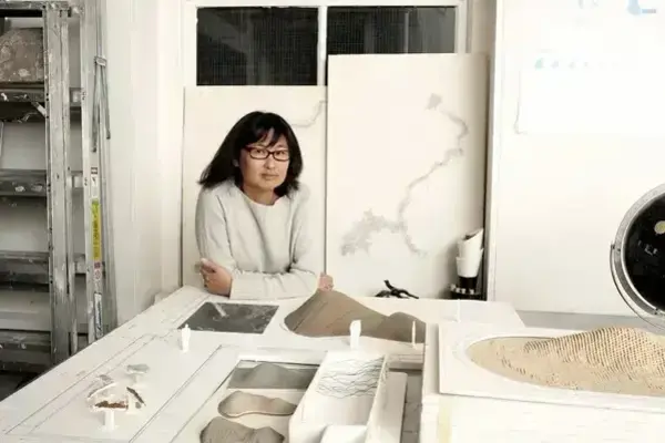 A woman with dark hair and glasses in a light sweater leans on a table displaying architectural models and plans.