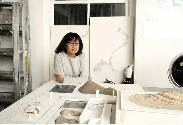 A woman with dark hair and glasses in a light sweater leans on a table displaying architectural models and plans.