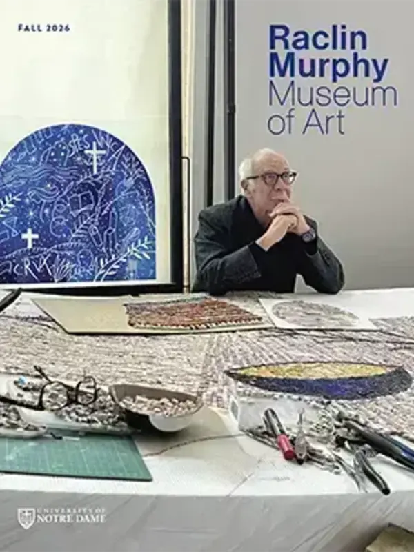 Artist in glasses observes intricate mosaic pieces on a table at Raclin Murphy Museum of Art. A blue arched artwork hangs behind him.