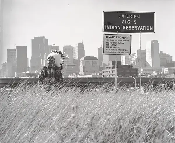 Man in headdress stands in grass before city skyline and reservation signs.